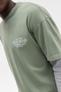 Person, der ein grünes BDG Jeans T-Shirt über einem grauen Langarmshirt trägt, zeigt nur das Brustlogo und den oberen Torso.
