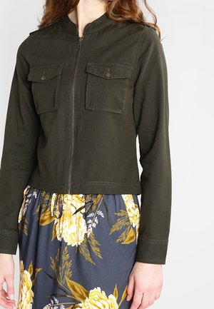 Vrouw draagt een donkergroene jas met rits en klepzakken en een bloemenrok met gele en bruine bloemen op een blauwe achtergrond.