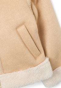 Veste beige clair avec une texture lisse et ressemblant à du daim, comportant une poche à l'avant et une doublure crémeuse douce et moelleuse au niveau de l'ourlet.