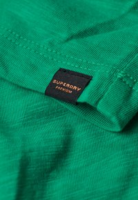 Groene katoenen T-shirt met een gestructureerd oppervlak, voorzien van een zwart label met de tekst "SUPERDRY PREMIUM" in oranje letters.