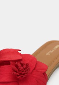 Κοντινό πλάνο ενός ταμπα πέδιλου με μεγάλη κόκκινη διακοσμητική υφασμάτινη ανθοδέσμη στο λουράκι πάνω σε λευκό φόντο.