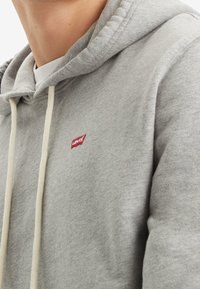 Šedá bavlněná mikina s kapucí se bílým šňůrkami, žebrovanými manžetami a malým červeným logem "Levi's" na hrudi. Měkká textura, pohodlný design.