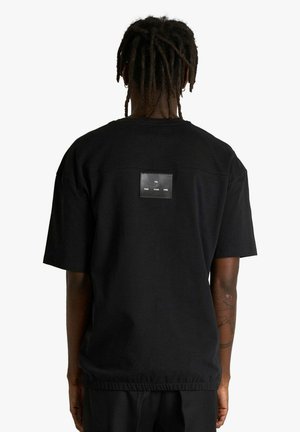 T-shirt nera in cotone con maniche corte e taglio dritto. Presenta una toppa rettangolare in pelle sul retro con testo in rilievo.