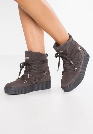 Jambes portant des bottines en daim gris foncé avec des semelles épaisses en caoutchouc noir, des lacets croisés et une doublure intérieure en fourrure douce.