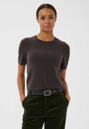 Femme aux cheveux foncés tressés portant un pull marron à manches courtes rentré dans un pantalon en velours côtelé vert avec une ceinture noire, mains dans les poches.