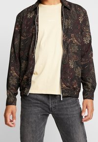 Man in een beige shirt met de tekst "Seoul", een zwart-rode paisley patroon zip-up jack en donkergrijze jeans tegen een witte achtergrond.