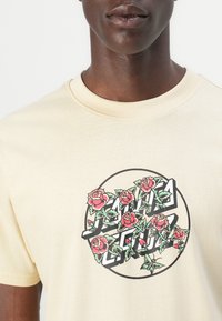 Short-sleeve cream T-shirt featuring a floral graphic with red roses and green stems, layered over black geometric shapes. Classic crew neck design.