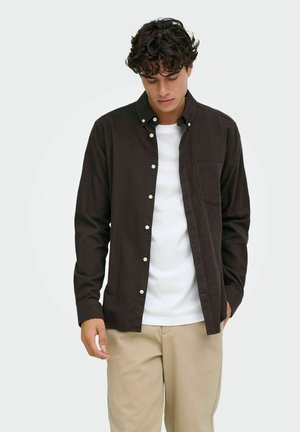 Jeune homme portant une chemise marron foncé déboutonnée sur un t-shirt blanc avec un pantalon beige, regardant vers le bas devant un fond uni.