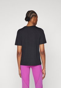 Schwarzes T-Shirt mit kurzen Ärmeln und rundem Halsausschnitt, glatter Textur und schlichtem Design. Getragen mit lila Leggings, die auffällige Nähte haben.