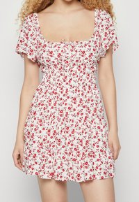 Blommig klänning med vit bakgrund och röda blommor, fyrkantig halsringning, korta svävande ärmar och en smockad midja.