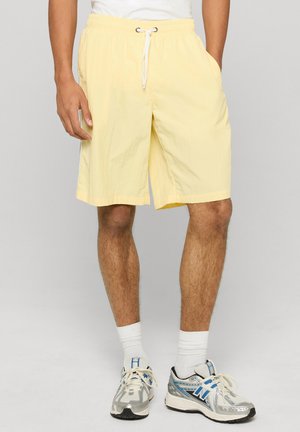 Homme portant un short à cordon de serrage jaune pâle, des chaussettes blanches et des chaussures de course argentées et bleues, debout devant un fond uni.