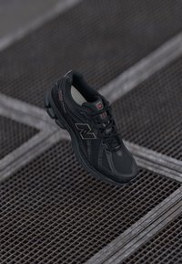 Musta New Balance jooksujalatse punaste aksentidega metallvõrgupinnal, nöörid seotud, nurga alt vaade, mis rõhutab külje logo ja tallakujundust.