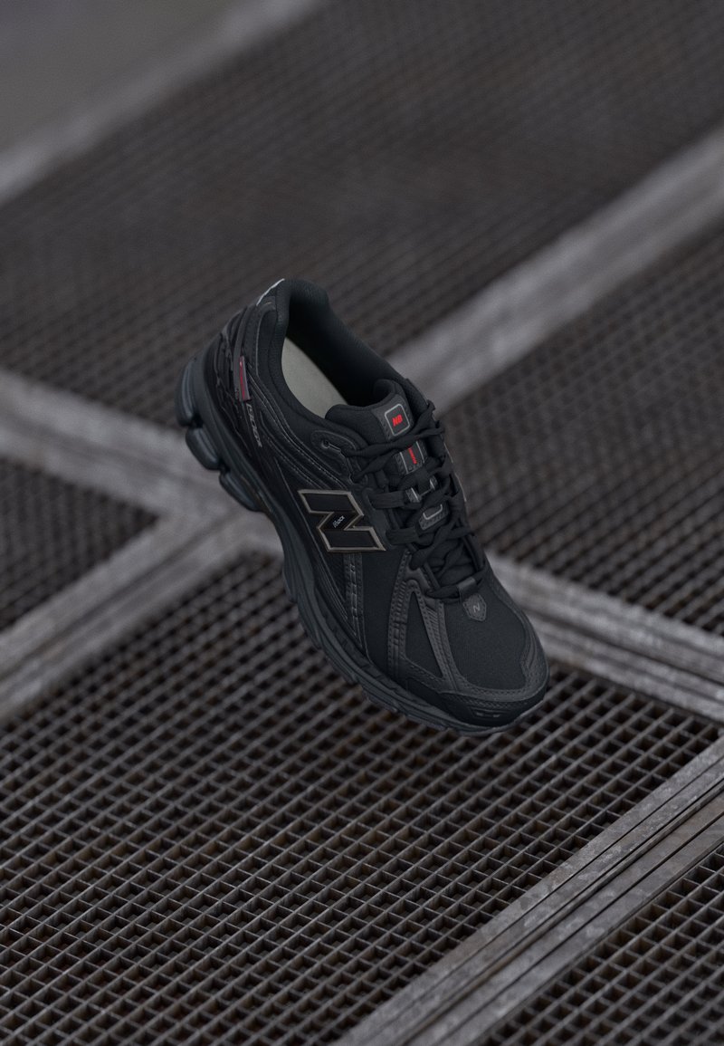 Musta New Balance jooksujalatse punaste aksentidega metallvõrgupinnal, nöörid seotud, nurga alt vaade, mis rõhutab külje logo ja tallakujundust.
