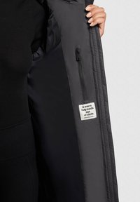 Interno di una giacca nera trapuntata con una tasca con zip. L'etichetta recita "Un caldo abbraccio fatto di tessuto." Tessuto di texture liscia e morbida.