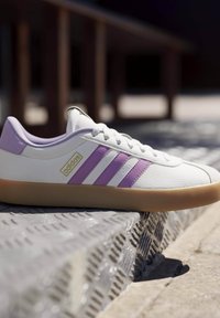 Zapatillas de cuero blancas con acentos morados claros y tres rayas moradas. Cuentan con una suela de goma y un logotipo dorado en el lateral.