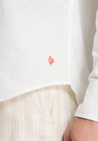 Weißes Leinenhemd mit einem gefalteten Saum und einem Detail aus orangefarbenem Korallenstickerei. Kombiniert mit hell gestreiften Stoffhosen, die eine lässige Passform zeigen.