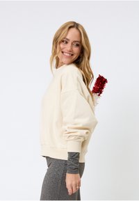 Sweat-shirt beige avec épaules tombantes et poignets côtelés, associé à des leggings gris pailletés ; un objet décoratif rouge et blanc apparaît à l'arrière.