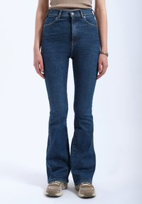 High-waisted flared jeans in dark blue denim with a smooth texture, featuring orange stitching and subtle pockets. Paired with beige sneakers.
