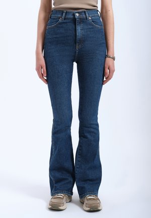 High-waisted flared jeans in dark blue denim with a smooth texture, featuring orange stitching and subtle pockets. Paired with beige sneakers.