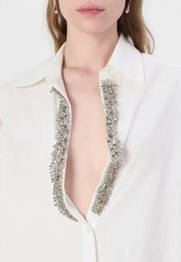 Bela majica z globokim V-izrezom, okrašena s srebrnimi beaded okraski ob ovratniku, kar ustvarja teksturiran kontrast do gladkega blaga.