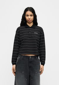 Νεαρή γυναίκα με μακριά κυματιστά μαλλιά, ντυμένη με μαύρο κοντό μπλουζάκι πόλο με λεπτές οριζόντιες ρίγες και φαρδιά σκούρα παντελόνια cargo.