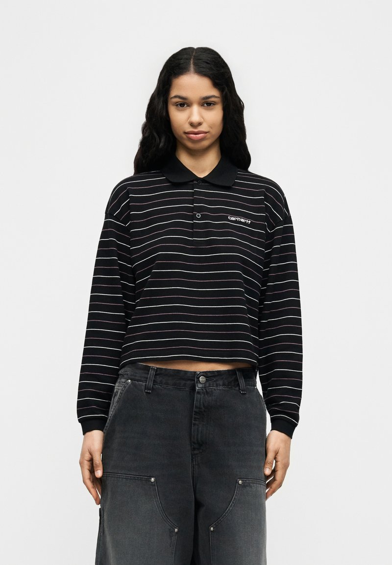 Jonge vrouw met lang golvend haar, gekleed in een zwart cropped polo shirt met lange mouwen en dunne horizontale strepen, gecombineerd met losse donkere cargobroeken.