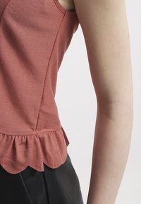 Top sin mangas de color rosa suave con un acabado texturizado, que presenta un dobladillo en ondas. El diseño tiene un ajuste entallado y se sitúa por encima de la cintura.