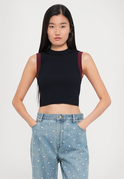 Priliehavý bezrukávový crop top z rebrovanej čiernej tkaniny s vínovými akcentmi na pleciach, doplnený o svetlo modré džínsy s bielymi bodkami.