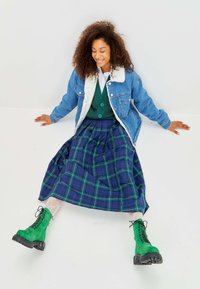 Jeansjack met witte bontkraag, groene sweatervest, blauwgroene geruite rok en groene veterschoenen tot de enkel op een witte achtergrond.