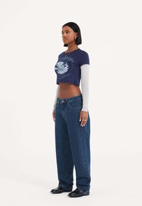 Maglietta cropped navy con design grafico sovrapposta a una camicia bianca a maniche lunghe, abbinata a jeans blu a vita alta e scollo largo.