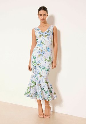 Frau steht in einem ärmellosen weißen Kleid mit blauem und grünem Blumenmuster und Rüschen am Saum, trägt hautfarbene High-Heels und goldenen Schmuck.