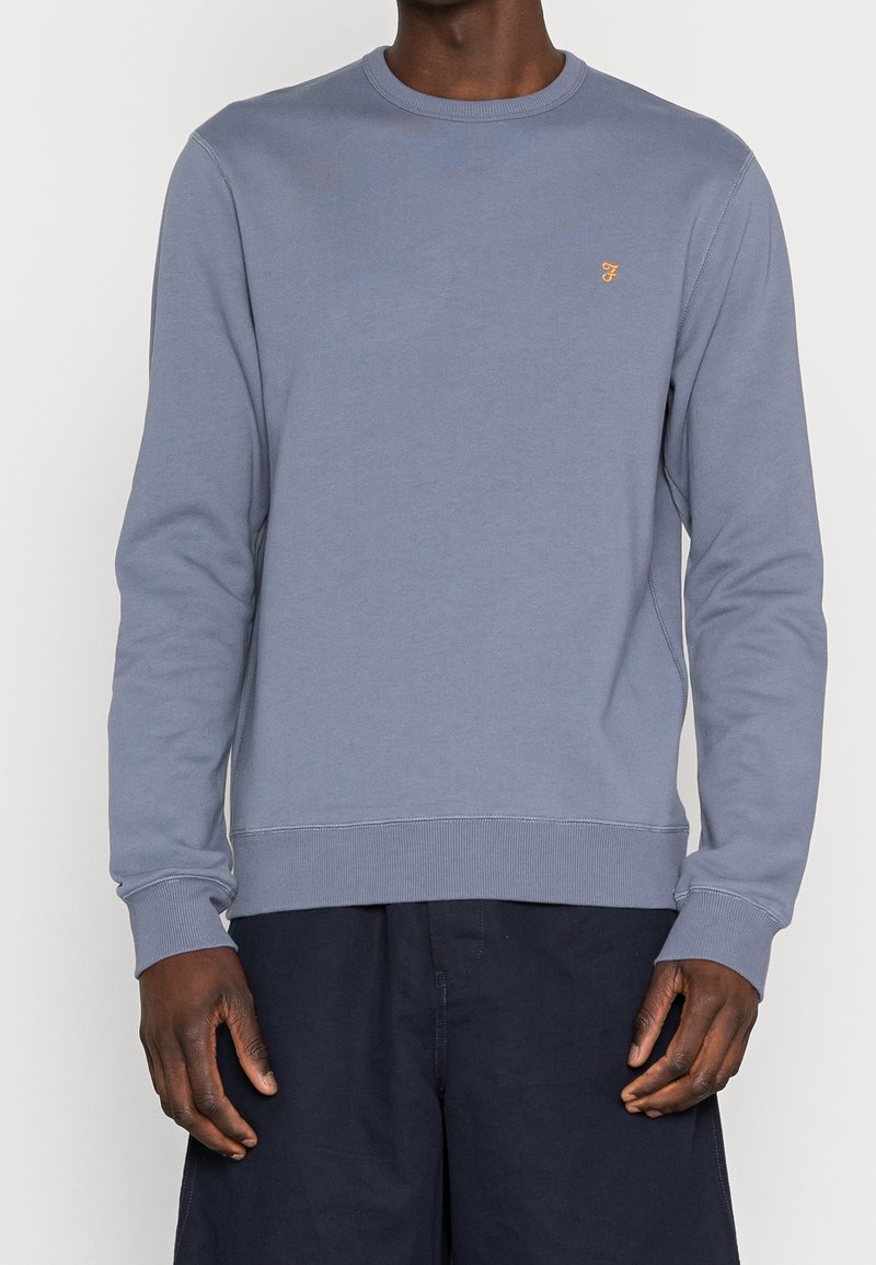Homme portant un sweat-shirt bleu clair à manches longues avec un petit logo orange brodé sur la poitrine et un pantalon foncé.