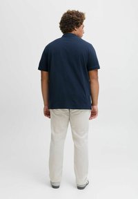 Dunkelblaues Polohemd mit einem Kragen, kurzen Ärmeln und hellbeigen, geraden Hosen. Graue Turnschuhe und eine strukturierte Frisur sind sichtbar.