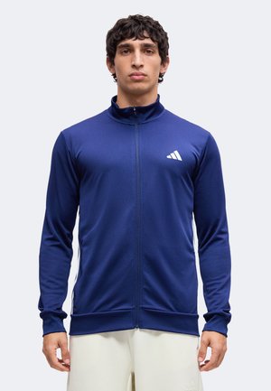 Mann trägt eine blaue Sportjacke mit Reißverschluss und weißem Logo auf der Brust sowie helle Hose, steht vor einfarbigem Hintergrund.