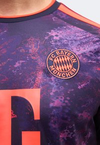 Φανέλα ποδοσφαίρου της FC Bayern München με μωβ και πορτοκαλί αφηρημένο σχέδιο και το έμβλημα του συλλόγου στο στήθος.