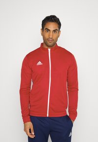 Czerwona sportowa kurtka z wysokim kołnierzem, białym zamkiem błyskawicznym i logo adidas na lewej piersi. Długie rękawy z dopasowanymi mankietami.