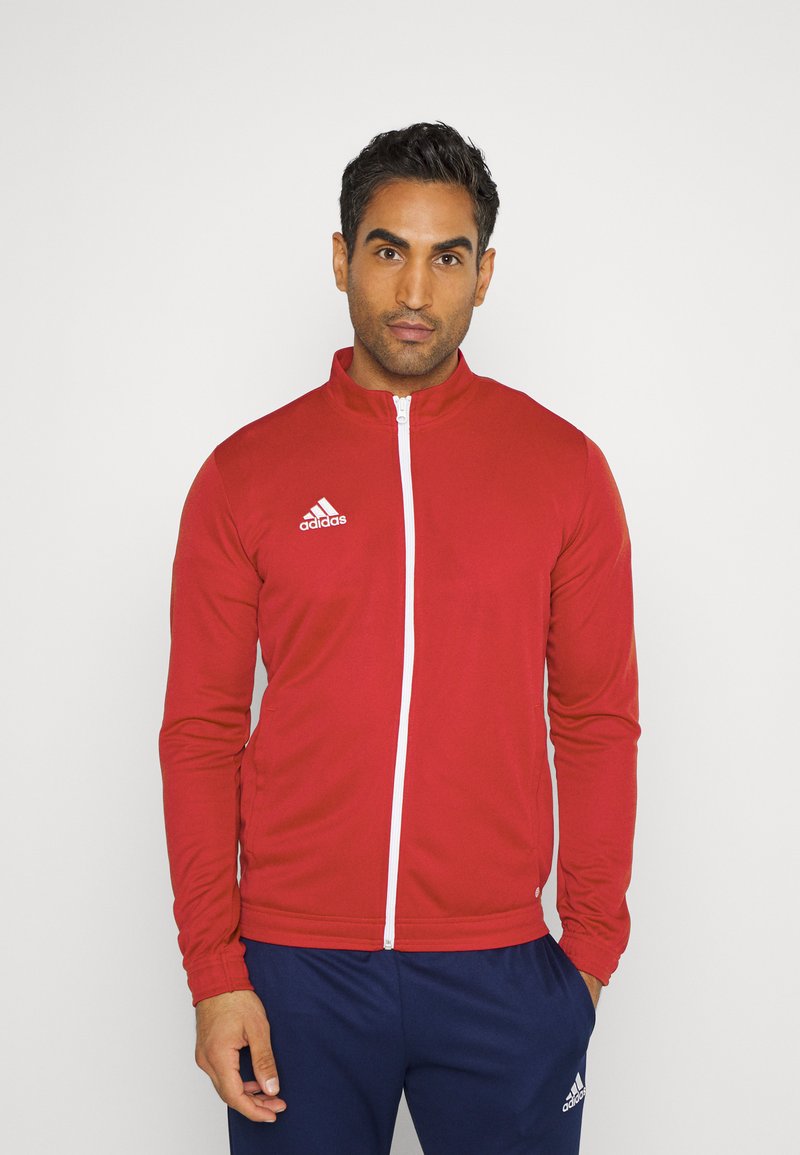 Czerwona sportowa kurtka z wysokim kołnierzem, białym zamkiem błyskawicznym i logo adidas na lewej piersi. Długie rękawy z dopasowanymi mankietami.