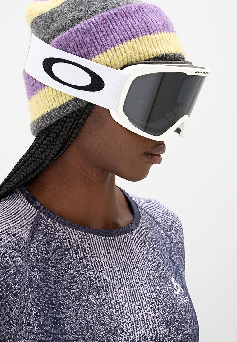 Maschera da sci con una montatura bianca e lenti scure, abbinata a un berretto a strisce grigio, giallo e viola. Maglietta sportiva con texture.