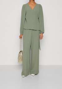Top avvolgente verde, texturizzato e a maniche lunghe abbinato a pantaloni larghi. Il modello tiene una borsa metallizzata con frange. Tacchi bianchi completano il look.