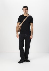 Camiseta negra, jeans oscuros holgados y zapatos negros. Una bolsa de mensajero beige cuelga cruzada sobre el cuerpo, con una correa simple y un detalle de hebilla.