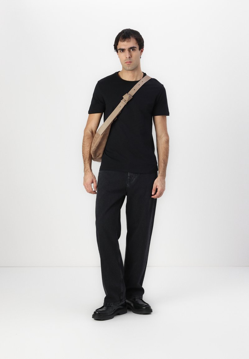 Camiseta negra, jeans oscuros holgados y zapatos negros. Una bolsa de mensajero beige cuelga cruzada sobre el cuerpo, con una correa simple y un detalle de hebilla.