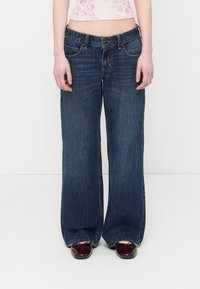 Γυναίκα που φορά φαρδιά σκούρα μπλε τζιν, μπορντό μπαλαρίνες και κοντό ροζ τοπ με λουλούδια, στέκεται μπροστά σε λευκό φόντο.