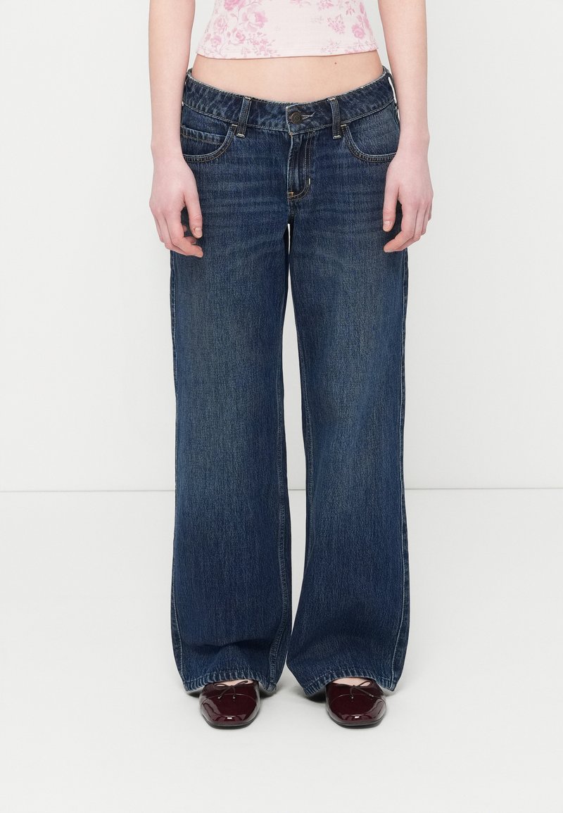 Γυναίκα που φορά φαρδιά σκούρα μπλε τζιν, μπορντό μπαλαρίνες και κοντό ροζ τοπ με λουλούδια, στέκεται μπροστά σε λευκό φόντο.