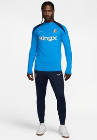 Maglietta sportiva a maniche lunghe blu con colletto alto e dettagli blu navy, caratterizzata da un grande logo bianco "BingX". Abbina a pantaloni sportivi navy e scarpe bianche.