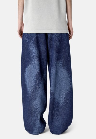 Person, die weit geschnittene, ausgewaschene dunkelblaue Jeans aus Denim trägt, von hinten vor neutralem Hintergrund gezeigt.