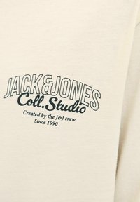 Beige tyg med svart text "JACK & JONES Coll. Studio Skapat av J&J-gänget Sedan 1990" tryckt på.