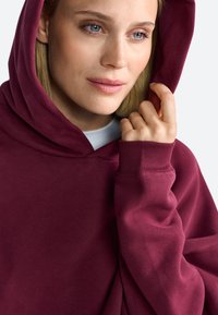 Bordeaux hoodie met een ruime capuchon, geribbelde manchetten en een zachte, gestructureerde stof. Het model houdt de capuchon vast en toont het ontspannen ontwerp.