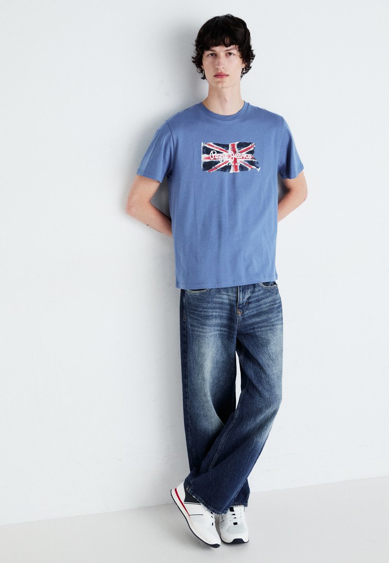 Blaues Baumwoll-T-Shirt mit Union Jack-Print, kombiniert mit locker sitzenden blauen Jeans und weißen Sneakern mit roten und schwarzen Akzenten.