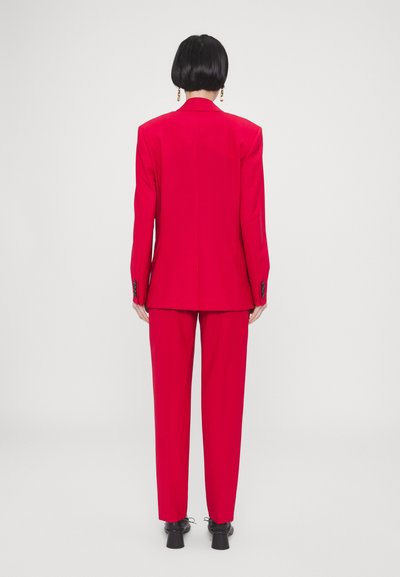 Costume sur mesure rouge avec blazer structuré et pantalons droits. Tissu lisse, détails de couture subtils, et chaussures noires.