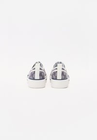 Hvite sneakers med et mønstret grått tekstilbakgrunn, flat gummisåle og en grepstropp på hælen. Elegant design med subtile detaljer.